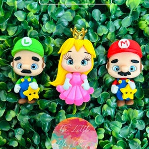 Puede incluir: Tres figuritas de arcilla hechas a mano de personajes de la serie de videojuegos Super Mario Bros. Las figuritas son de Mario, Luigi y la princesa Peach. Mario lleva su atuendo rojo y azul característico, Luigi lleva su atuendo verde y azul característico y la princesa Peach lleva su vestido rosa y corona característicos. Las figuritas están todas de pie sobre un fondo verde.