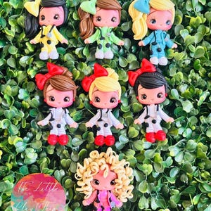 May include: A collection of handmade clay figurines of dolls with various hair colors and bows. Each doll wears a different colored outfit, including yellow, green, blue, and white. The dolls have detailed facial features and are set against a green leafy background. The Little Clay Shop.