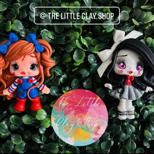 Puede incluir: Dos figuras de arcilla hechas a mano de muñecas. Una muñeca tiene pelo rojo, lazos azules y usa un mono azul. La otra muñeca tiene pelo negro, un lazo blanco y una falda gris. El texto "@ THE LITTLE CLAY SHOP" está en la parte superior.