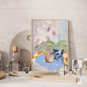 Puede incluir: Una pintura de acuarela enmarcada de orquídeas blancas con acentos rosados, un jarrón azul y un recipiente cilíndrico marrón. La pintura descansa sobre una mesa con otros elementos decorativos, incluyendo tablas de cortar de madera y recipientes de vidrio.
