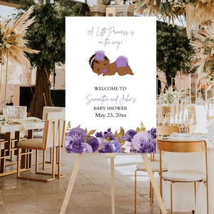 Può includere: Un cartello di benvenuto per un baby shower su un cavalletto di legno. Il cartello recita "A Little Princess is on the way!" e "Welcome to Samantha and John's Baby Shower May 23, 20xx." Il cartello ha un bordo floreale con fiori viola.