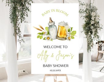 May include: A white sign with green and yellow floral accents. The sign reads "Baby in Bloom" and features a baby bottle and a beer mug. The text below reads "Welcome to Ally & Jason's Baby Shower 05.22.20XX".