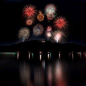 May include: A nighttime scene features a display of colorful fireworks bursting in the sky. The fireworks are reflected in the dark water below, with lights along the shoreline. The fireworks are red, white, gold, and green.