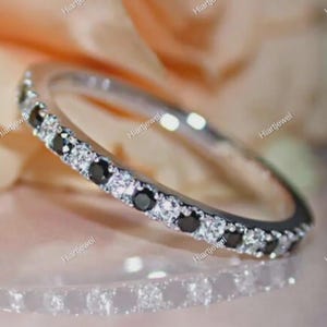 May include: A silver band ring with alternating black and clear gemstones. The ring is set against a soft, blurred background, highlighting the sparkle of the stones. The ring is a delicate piece of jewelry.