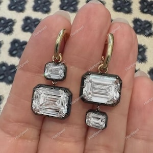 以下が含まれることがあります： エメラルドカットのクリアな宝石が付いた、金のフープピアス。イヤリングには、宝石の周りに黒い縁取りがあります。イヤリングは、模様のある背景に対して表示されます。