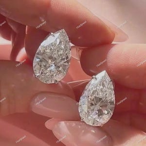 May include: Two pear-shaped, colorless, faceted gemstones set in silver-toned metal prongs. Each stone features a pointed top and a rounded base, displaying high clarity and brilliance with multiple internal light reflections throughout the facets.