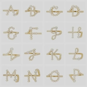 Könnte beinhalten: Eine Sammlung goldfarbener Ringe, die jeweils als ein Buchstabe des Alphabets gestaltet sind. Die Buchstaben werden aus kleinen, runden, klaren Edelsteinen gebildet. Die Ringe werden vor einem hellen Hintergrund präsentiert.