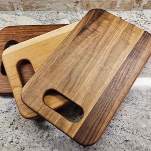 May include: Three wooden cutting boards with handles. The top board is made of walnut wood and has a darker brown color. The middle board is made of maple wood and has a lighter brown color. The bottom board is made of cherry wood and has a reddish brown color.