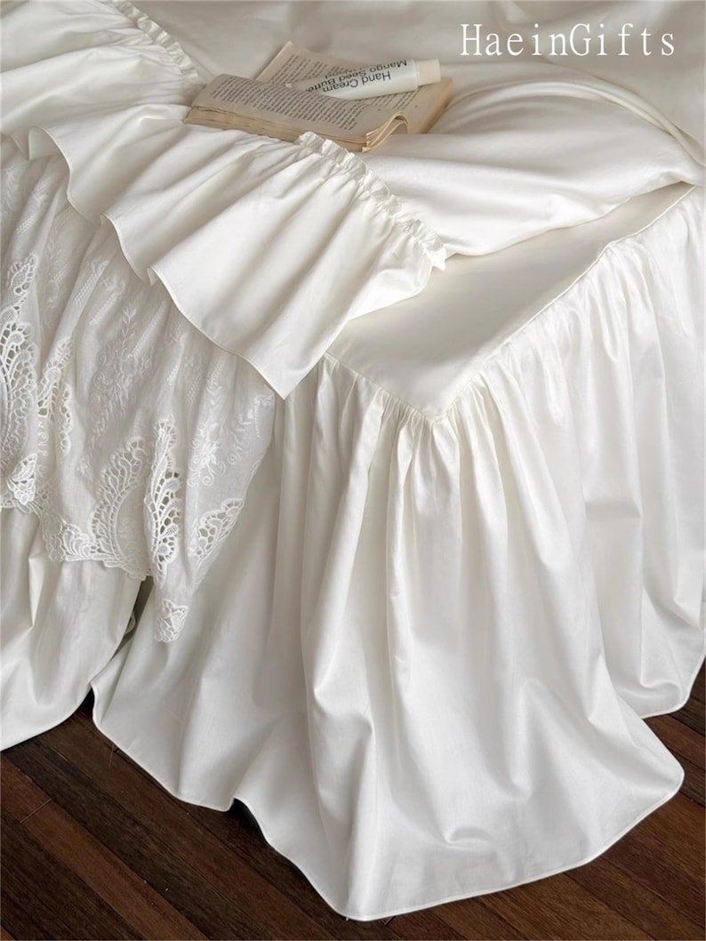 May include: White bedding set with ruffled edges and a lace detail. A book and a tube of hand cream rest on the bed. The bedding has a smooth, satin-like texture, creating a luxurious look. The brand name "HaeinGifts" is visible in the top right corner.