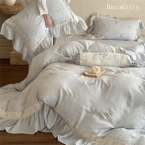 May include: A light blue bedspread with a white lace trim and embroidered floral designs. The bedspread is made of a soft, lightweight fabric and has a ruffled edge. The bedspread is paired with matching pillowcases and a decorative pillow with a lace trim and the text "London Rose".