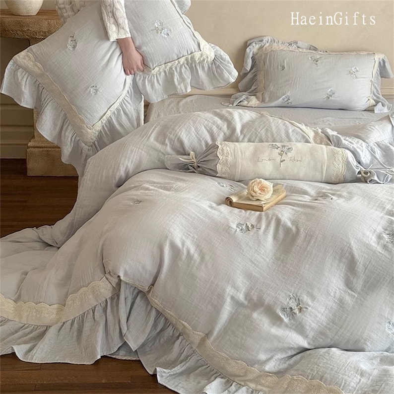 May include: A light blue bedspread with a white lace trim and embroidered floral designs. The bedspread is made of a soft, lightweight fabric and has a ruffled edge. The bedspread is paired with matching pillowcases and a decorative pillow with a lace trim and the text "London Rose".