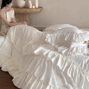 May include: Cream-colored bedding set with ruffled edges and lace detailing. The set includes a bed skirt, pillows, and a duvet cover. The image also shows a woman sitting on the bed, holding a book. The text "HaeinGifts" is visible in the top right corner.