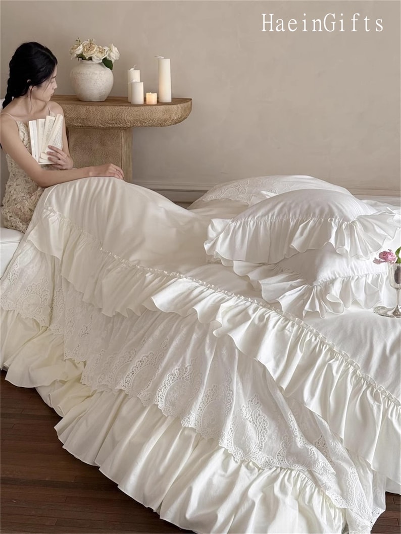 May include: Cream-colored bedding set with ruffled edges and lace detailing. The set includes a bed skirt, pillows, and a duvet cover. The image also shows a woman sitting on the bed, holding a book. The text "HaeinGifts" is visible in the top right corner.