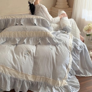 May include: A light blue bedspread with white lace trim and ruffles. The bedspread has a delicate floral embroidery pattern.
