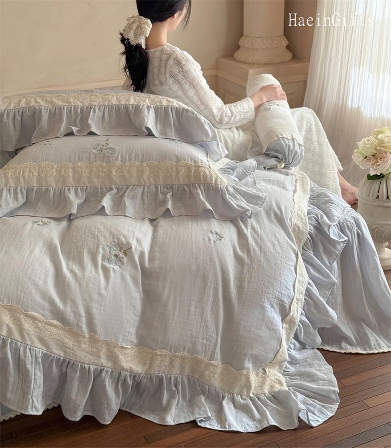 May include: A light blue bedspread with white lace trim and ruffles. The bedspread has a delicate floral embroidery pattern.