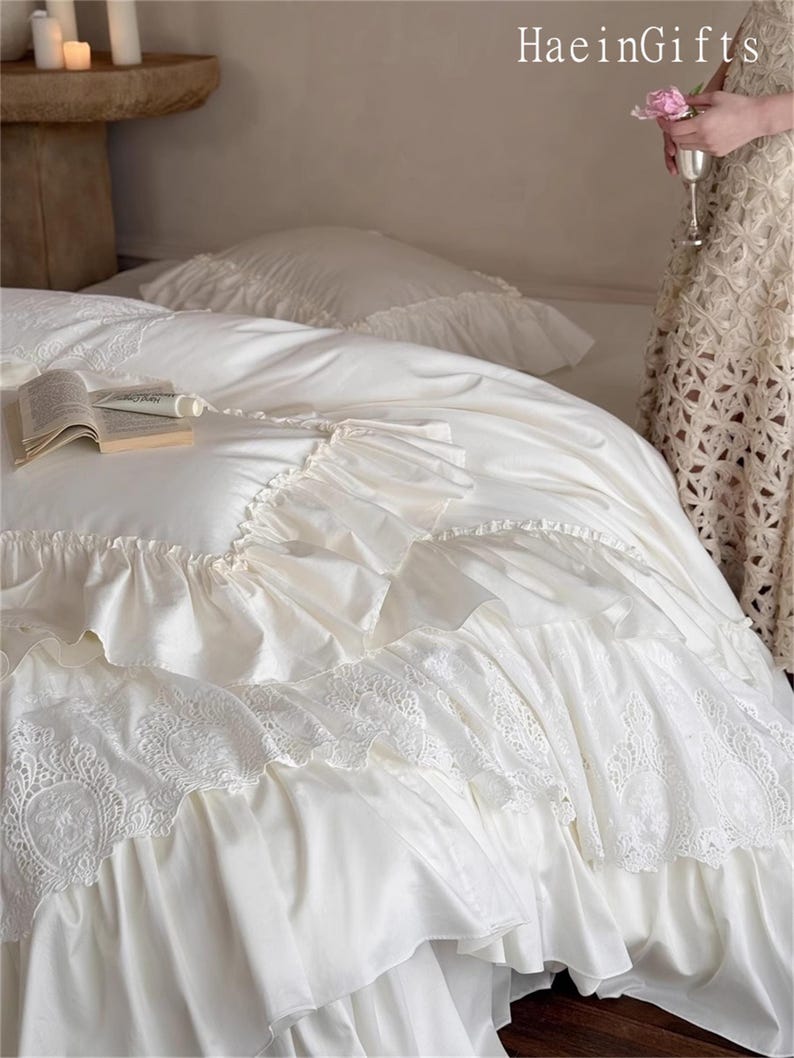 May include: Ivory-colored bedding set with ruffled edges and lace detailing. The set includes a duvet cover and pillowcases. A book rests on the bed. The text "HaeinGifts" is visible in the upper right corner.