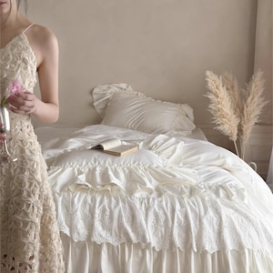 May include: Cream-colored bedding set with ruffled and lace details. The bed is made with a white comforter, pillow, and a decorative bed skirt. A book rests on the bed, and the text "HaeinGifts" is visible in the upper right corner.