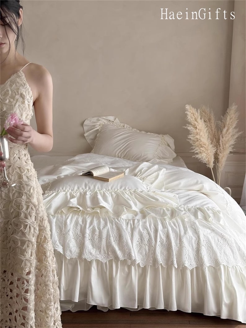 May include: Cream-colored bedding set with ruffled and lace details. The bed is made with a white comforter, pillow, and a decorative bed skirt. A book rests on the bed, and the text "HaeinGifts" is visible in the upper right corner.