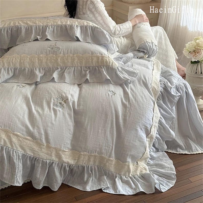 May include: A light blue bedspread with white lace trim and ruffles. The bedspread is embroidered with small floral designs. The bedspread is on a bed with white pillows and a white throw pillow.