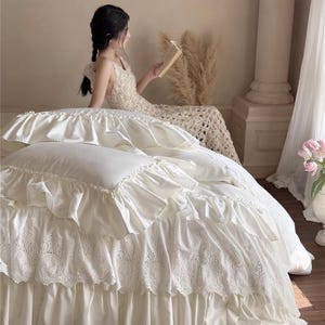 May include: Cream-colored bedding set with ruffled edges and intricate lace detailing. The bedspread features multiple layers of fabric, creating a textured and elegant look. The image includes the text "HaeinGifts".