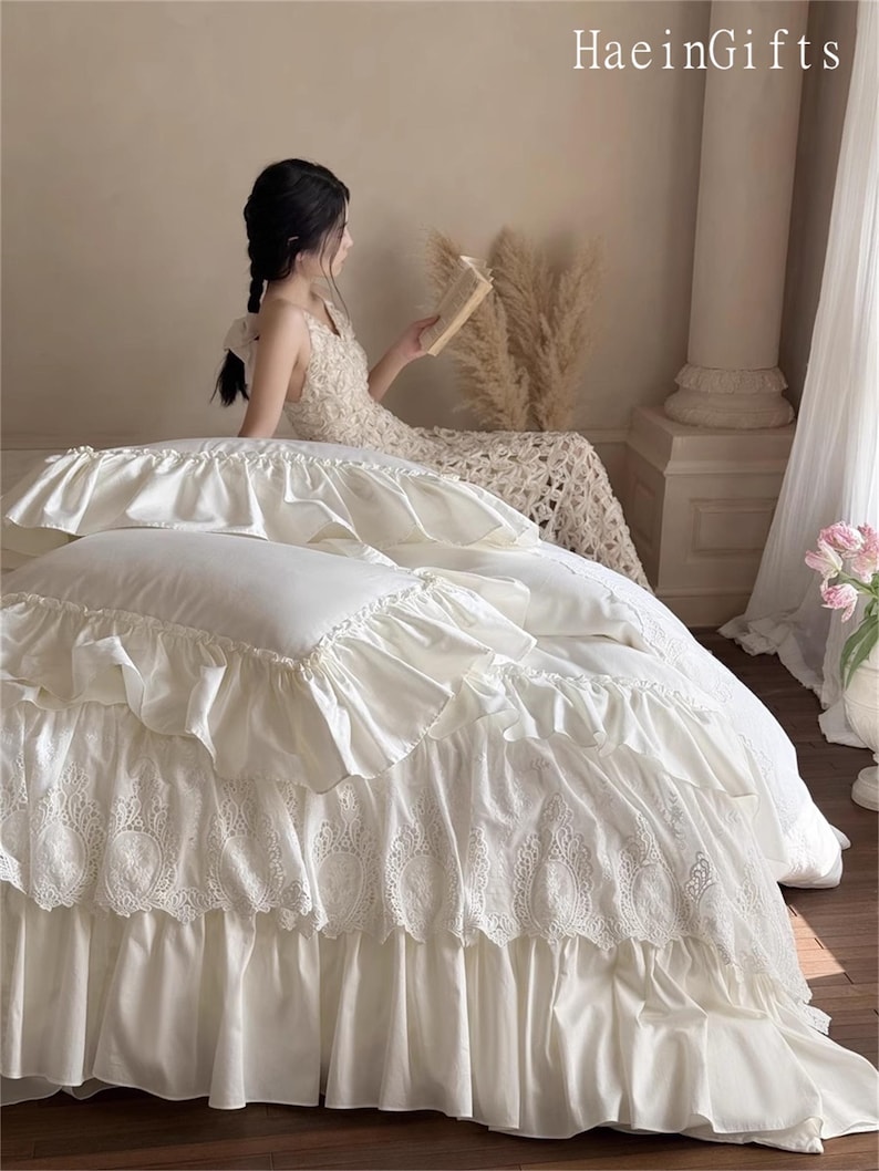 May include: Cream-colored bedding set with ruffled edges and intricate lace detailing. The bedspread features multiple layers of fabric, creating a textured and elegant look. The image includes the text "HaeinGifts".