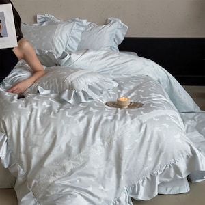 May include: Light blue bedding set with a butterfly pattern. The set includes a duvet cover, pillowcases, and decorative trim. The bedding has a ruffled edge and a soft, silky appearance. A lit candle sits on a small plate.