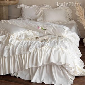 May include: A luxurious white bedding set with ruffled edges and lace detailing. The set includes pillows, a duvet, and a bed skirt. A small pink flower and a glass are on the bed. The text "HaeinGifts" is visible in the upper right corner.