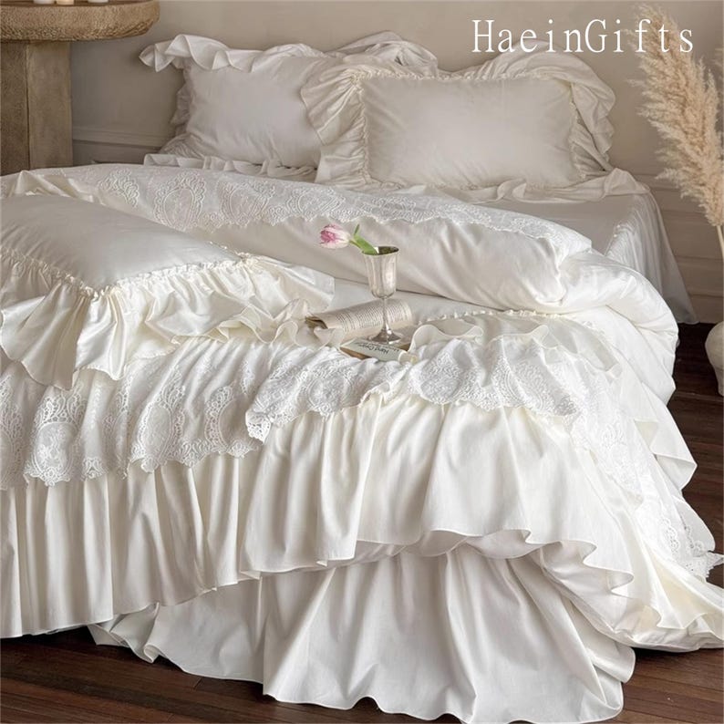 May include: A luxurious white bedding set with ruffled edges and lace detailing. The set includes pillows, a duvet, and a bed skirt. A small pink flower and a glass are on the bed. The text "HaeinGifts" is visible in the upper right corner.