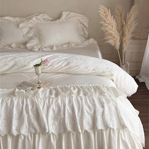 May include: A white bed set with ruffled pillowcases and a decorative bed skirt. The bedding features intricate lace detailing and a soft, flowing design. A small vase with a pink flower and a book are on the bed.