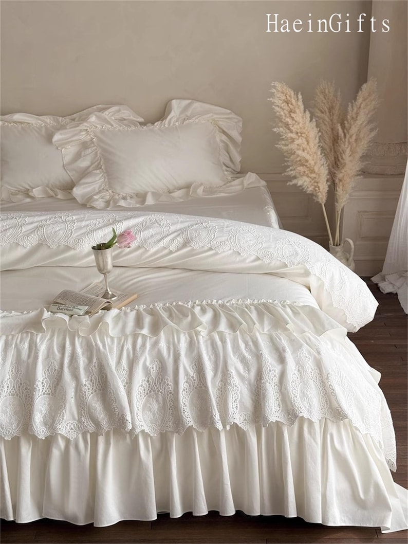 May include: A white bed set with ruffled pillowcases and a decorative bed skirt. The bedding features intricate lace detailing and a soft, flowing design. A small vase with a pink flower and a book are on the bed.