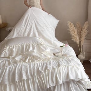 May include: Ivory-colored bedding set with ruffled edges and lace detailing. The set includes a comforter, pillow, and bed skirt. The image also shows a person holding the comforter. The text "HaeinGifts" is visible in the upper right corner.