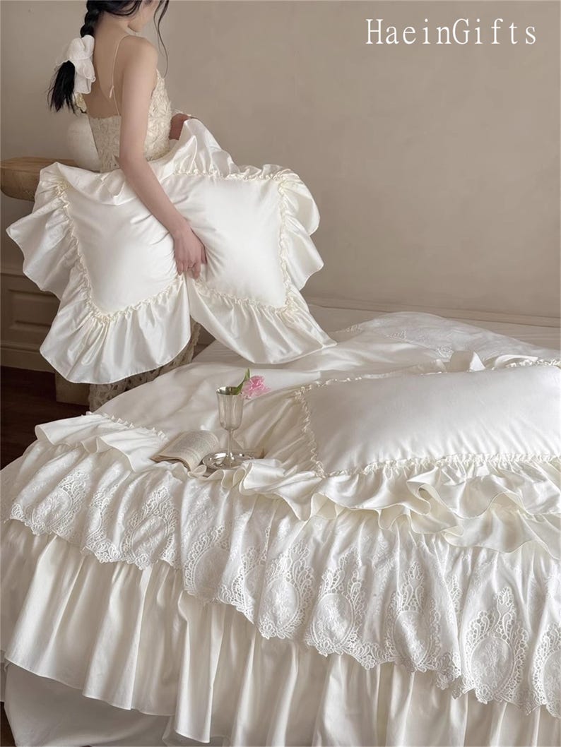 May include: Ivory-colored bedding set with ruffled edges and lace detailing. The set includes a duvet cover and pillowcases. The image shows a bed with the bedding arranged, with the text "HaeinGifts" in the upper right corner.