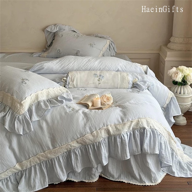May include: A light blue bedspread with white lace trim and ruffles. The bedspread is made of a soft, textured fabric and has a subtle striped pattern. The bed is decorated with white pillows and a decorative pillow with a floral design.