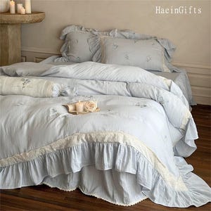 May include: A light blue bed set with a white lace trim and floral embroidery. The bedding features a ruffled design and a pillow with the text "London Sky".