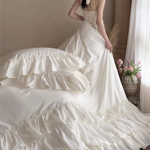 May include: Ivory-colored bedding set with ruffled edges and lace detailing. The set includes a fitted sheet, pillowcases, and a duvet cover. The image also shows a woman wearing a white dress. The text "HaeinGifts" is visible in the upper right corner.