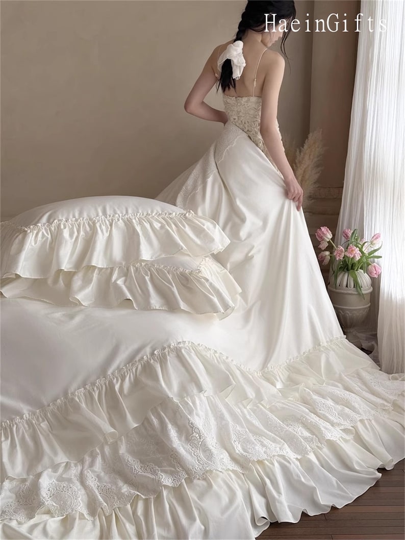 May include: Ivory-colored bedding set with ruffled edges and lace detailing. The set includes a fitted sheet, pillowcases, and a duvet cover. The image also shows a woman wearing a white dress. The text "HaeinGifts" is visible in the upper right corner.