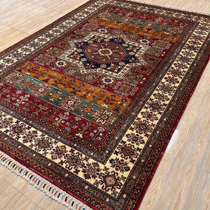 Puede incluir: Una alfombra persa tradicional con un patrón geométrico colorido. La alfombra tiene un esquema de color rojo, naranja, azul, verde y crema. La alfombra tiene un borde de flecos.