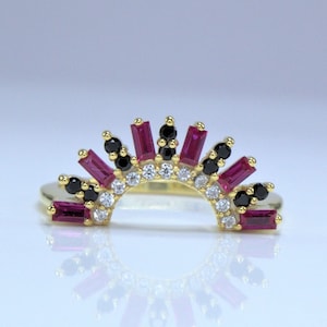 May include: A gold-colored ring with a sunburst design. The ring features a series of rectangular, ruby-colored stones, interspersed with black and white stones. The ring is set against a white background.