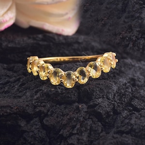 Op de afbeelding: Een gouden ring met een rij ovale gele edelstenen. De ring is geplaatst tegen een donkere, getextureerde achtergrond, die de heldere gele stenen benadrukt. De ring heeft een eenvoudige, gepolijste gouden band.