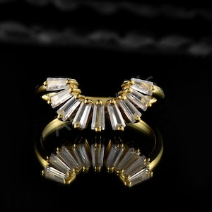 Op de afbeelding: Een goudkleurige ring met een waaiervormig ontwerp van heldere, rechthoekige stenen. De ring staat tegen een zwarte achtergrond, met een weerspiegeling eronder. Het ontwerp is symmetrisch en elegant.