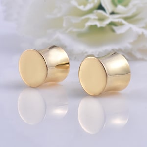 Puede incluir: Dos tapones para los oídos dorados pulidos con forma cilíndrica y una cara plana y circular. Los tapones se muestran sobre una superficie reflectante, con una flor blanca borrosa en el fondo. Los tapones son de color dorado brillante.