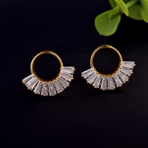Peut inclure: Une paire de boucles d'oreilles dorées composées d'un anneau circulaire orné d'un éventail de cristaux transparents taille baguette sur la moitié inférieure. Le design crée un effet rayon de soleil avec des pierres scintillantes sur métal.
