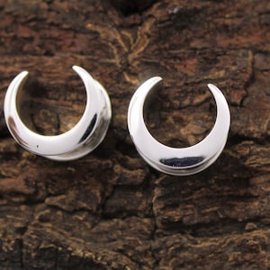 May include: Two silver crescent moon shaped ear gauges. The gauges are made of metal and have a smooth, polished finish.