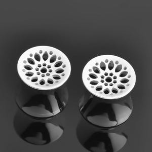 Könnte beinhalten: Zwei weiße, kreisförmige Ohrringe mit floralem Design. Jeder Ohrring hat kleine, ovale Aussparungen um ein zentrales Loch. Die Ohrringe liegen auf einer dunklen, reflektierenden Oberfläche.