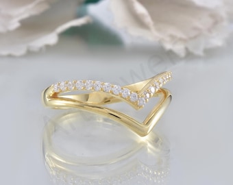 May include: A gold-colored ring with a V-shaped design. The top band is adorned with small, clear gemstones. The ring is set against a soft, blurred background, highlighting its elegant and modern style.