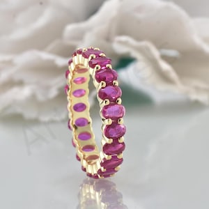 May include: A gold ring with a continuous band of oval-cut, vibrant pink gemstones. The gemstones are set closely together, creating a full circle of colour. The ring's gold band provides a warm contrast to the bright gemstones.