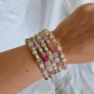 May include: A stack of beaded bracelets in shades of pink, white, and gold. The bracelets feature a variety of bead shapes and sizes, including round, faceted, and oval beads. The bracelets are worn on a wrist against a white background.