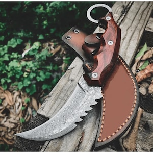 Op de afbeelding: Een gebogen karambitmes met een patroon op het lemmet en een houten handvat. Het mes zit gedeeltelijk in een bruin leren holster. De handgreep is donkerbruin en vastgezet met zilveren klinknagels.