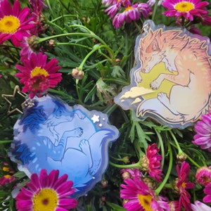 May include: Two clear acrylic charms featuring a blue wolf and an orange wolf, both sleeping on clouds with stars. The charms are hanging from a chain and are surrounded by pink flowers.
