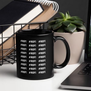 Puede incluir: Taza de café negra con texto blanco que dice '#REF!' repetido varias veces. La taza está sobre una superficie blanca junto a un portátil y una planta en maceta.